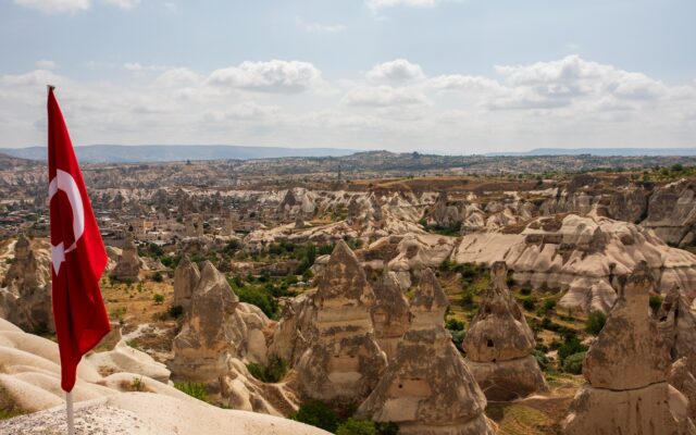 I camini delle fate della Cappadocia: concrezioni di roccia che si ergono dal terreno. In primo piano, a sinistra, una bandiera della Turchia, rossa con una stella bianca.